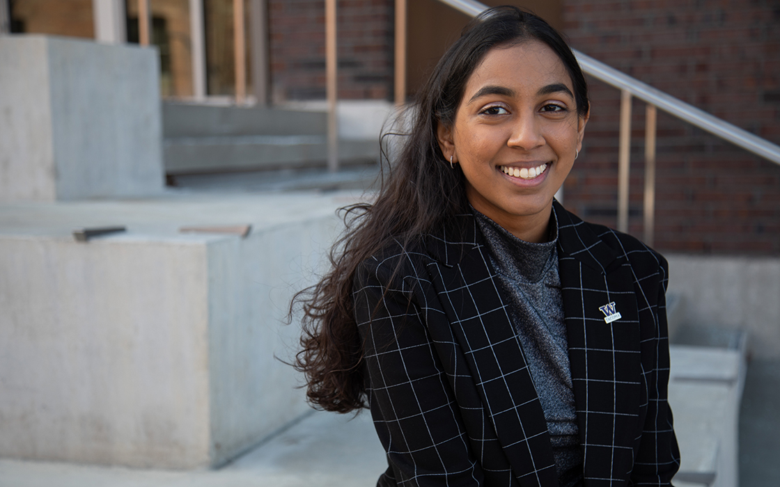 UW Tacoma student Angel Reddy sits on the bottom step of a series of concrete steps. She is wearing a red checkered coat. There is a purple and gold UW Tacoma pin on her lapel. There are bricks and a handrail in the background. Reddy is smiling and has long black hair.