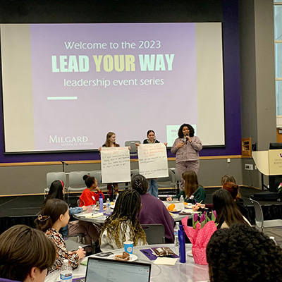 Three people are onstage. Two of the people are holding posters in their hands. One is speaking. In the background is the projected image of a logo for Lead Your Way.