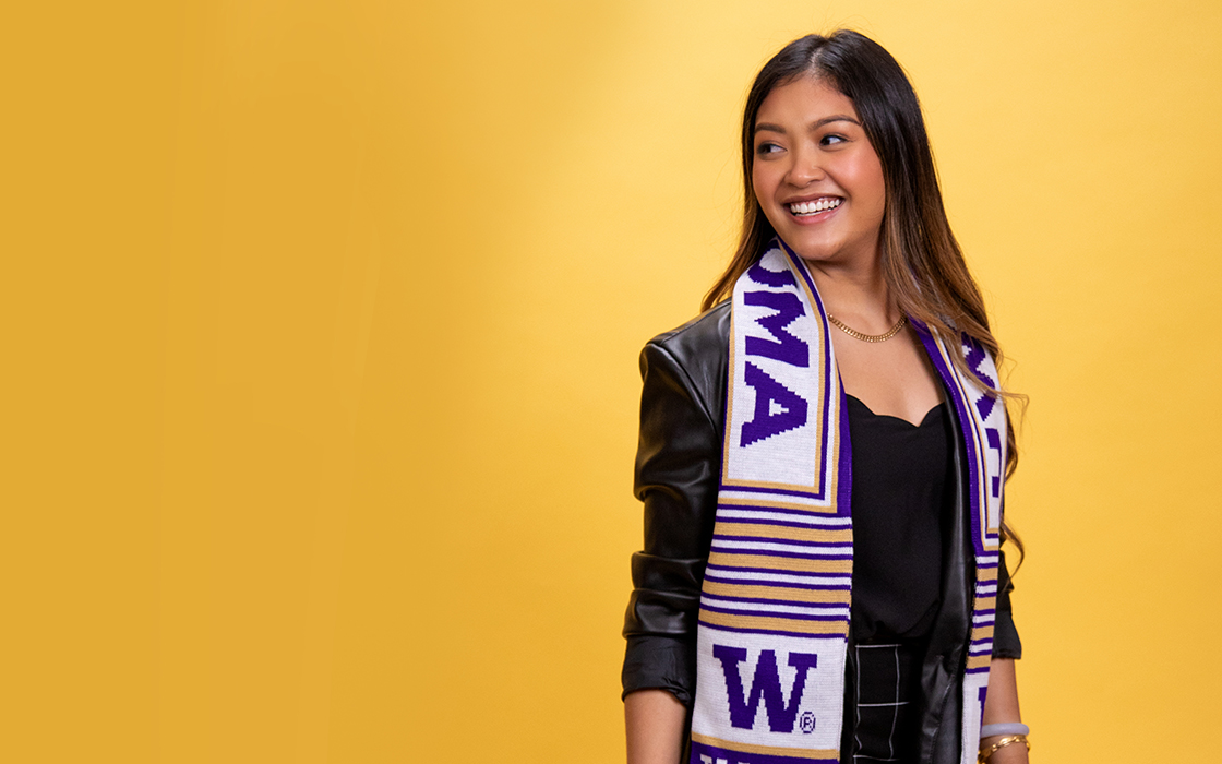 UW Tacoma alumna Victoria Nuon stands against a gold background. She has long brown hair and is wearing a purple, gold and white UW Tacoma scarf. She has a dark colored blouse on.