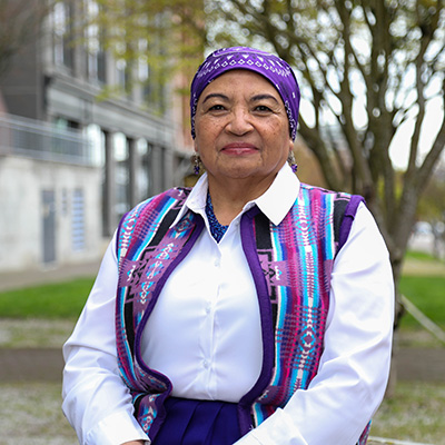 Ada McDaniel sits on a bench. There are trees behind her. She is wearing a purple headwrap with symbols sewn in. She is also wearing a purple vest with symbols sewn in. McDaniel has on a white undershirt.