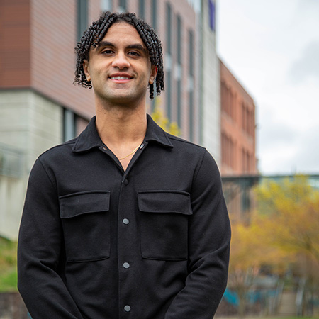 UW Tacoma student Martin Shehata stands in front of the Tioga library. There are trees and the library in the background. Shehata has short, dark hair and is smiling. He is wearing a black button up shirt.