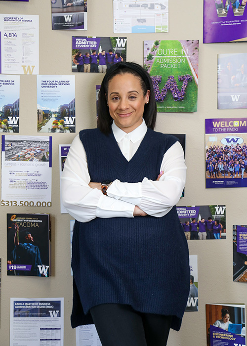 UW Tacoma staff member Navia Winderling leans against a wall. She is wearing a purple sweater vest over a white shirt. She has on a dark colored skirt. Behind her are a handful of UW Tacoma brochures that are attached to the wall.