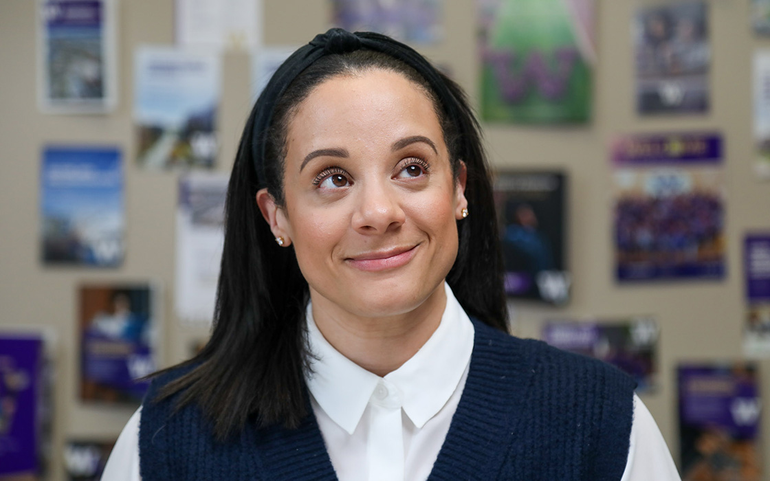 Headshot of staff member Navia Winderling. Navia has long, dark hair. She is wearing a purple sweater vest under a white shirt. There are a handful of UW Tacoma promotional brochures on the wall behind her.