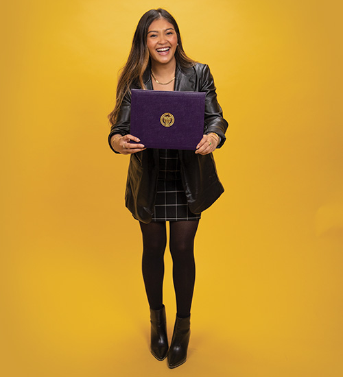 UW Tacoma alumna Victoria Nuon stands against a gold background. She is holding a UW degree cover between her hands. Nuon is smiling. She has long brown hair and is wearing a dark colored blouse and skirt.