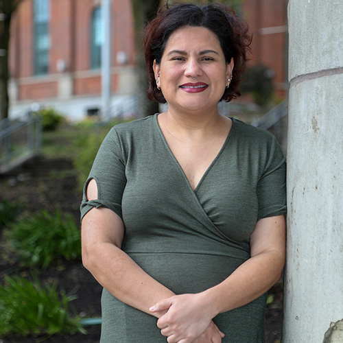 Recent UW Tacoma Master of Nursing graduate Juana Gallegos stands against a cement column. She is wearing a brown dress and has her arms crossed in front of her. She has short, dark hair. There are bushes and a building in the background.