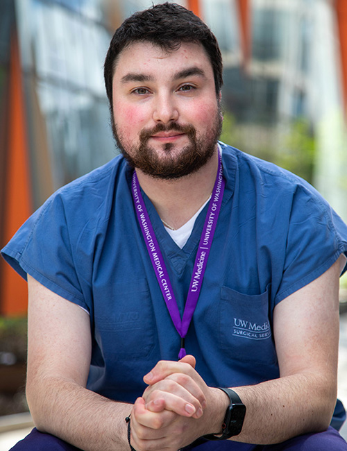 UW Tacoma student Lucas Bjorkheim sits on bench. He is wearing blue medical scrubs and has a name tag hanging around his neck. Lucas has short black hair and a black beard. There is an out of focus series of building windows behind him.