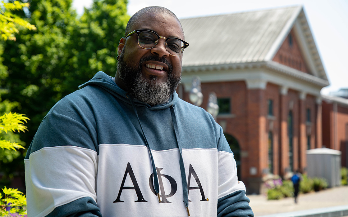 Headshot of UW Tacoma alumnus Jeff Dade. Dade is wearing a green and white sweatshirt with Greek (fraternity) letters on it. There is a tree with green leaves in the background as well as the Snoqualmie Powerhouse building which is made of red brick.