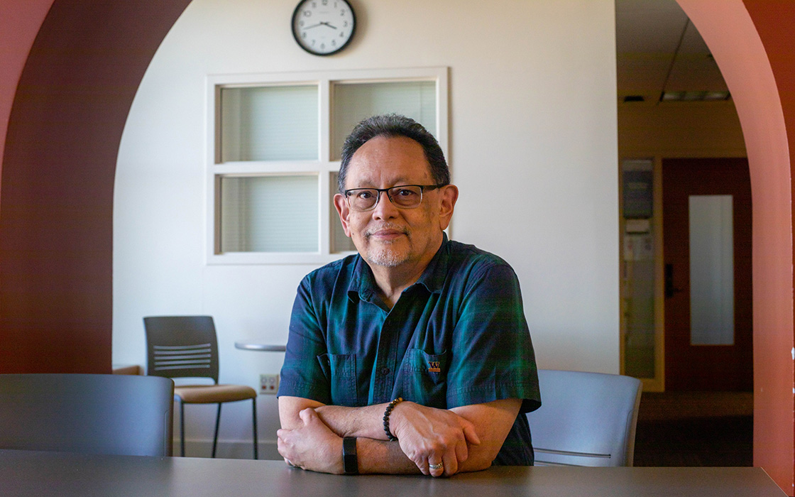 UW Tacoma faculty member Ronnie San Nicolas sits at a desk, his arms crossed in front of him on a table. There is a white wall with a window and a clock behind him. San Nicolas is wearing a dark colored, stripped shirt. He has glasses and a goatee.