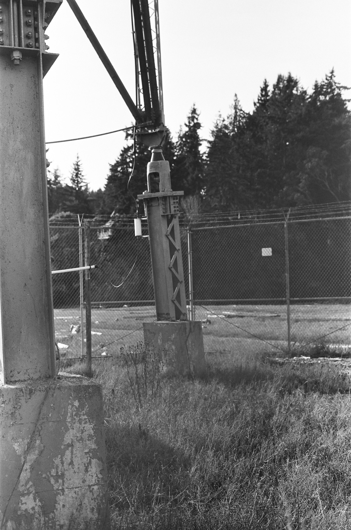 PICTURED: A black and white photo of the legs of a fenced in radio communication tower. It is enclosed by chainlink fence. Trees are in the background.