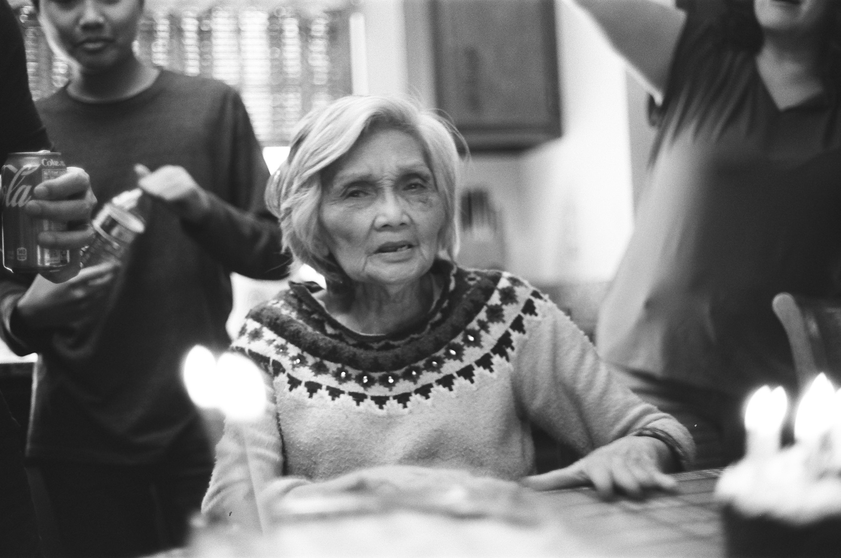 PICTURED: A black and white portrait of the photographer's grandmother. She sits while other people stand beside and behind her, partially out of frame. Cakes with candles are blurry in the foreground.
