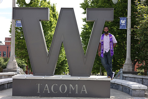 UW Tacoma alumnus Joe Davis stands next to the large steel W on campus. Davis is wearing blue jeans, a blue plaid shirt and a purple stole around his neck that says "veteran." Davis has short, dark hair. There are trees behind him and a light pole