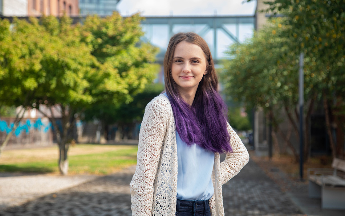 UW Tacoma student Holly Wetzel stands on the Prairie Line Trail on campus. Her left arm is on her him and her right arm is straight by her side. Wetzel is wearing a long white coat with a white undershirt. Her hair is brown with purple tips. There are trees and a skybridge in the background.