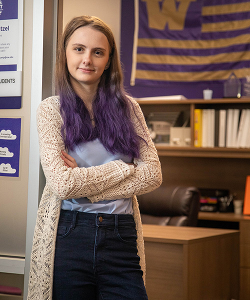 UW Tacoma student Holly Wetzel stands in the doorway of her ASUWT office. There is a purple and gold UW flag in the background as well as books, chair and a desk. Wetzel is wearing a long white coat. white shirt and blue jeans. Her hair is brown with purple tips.