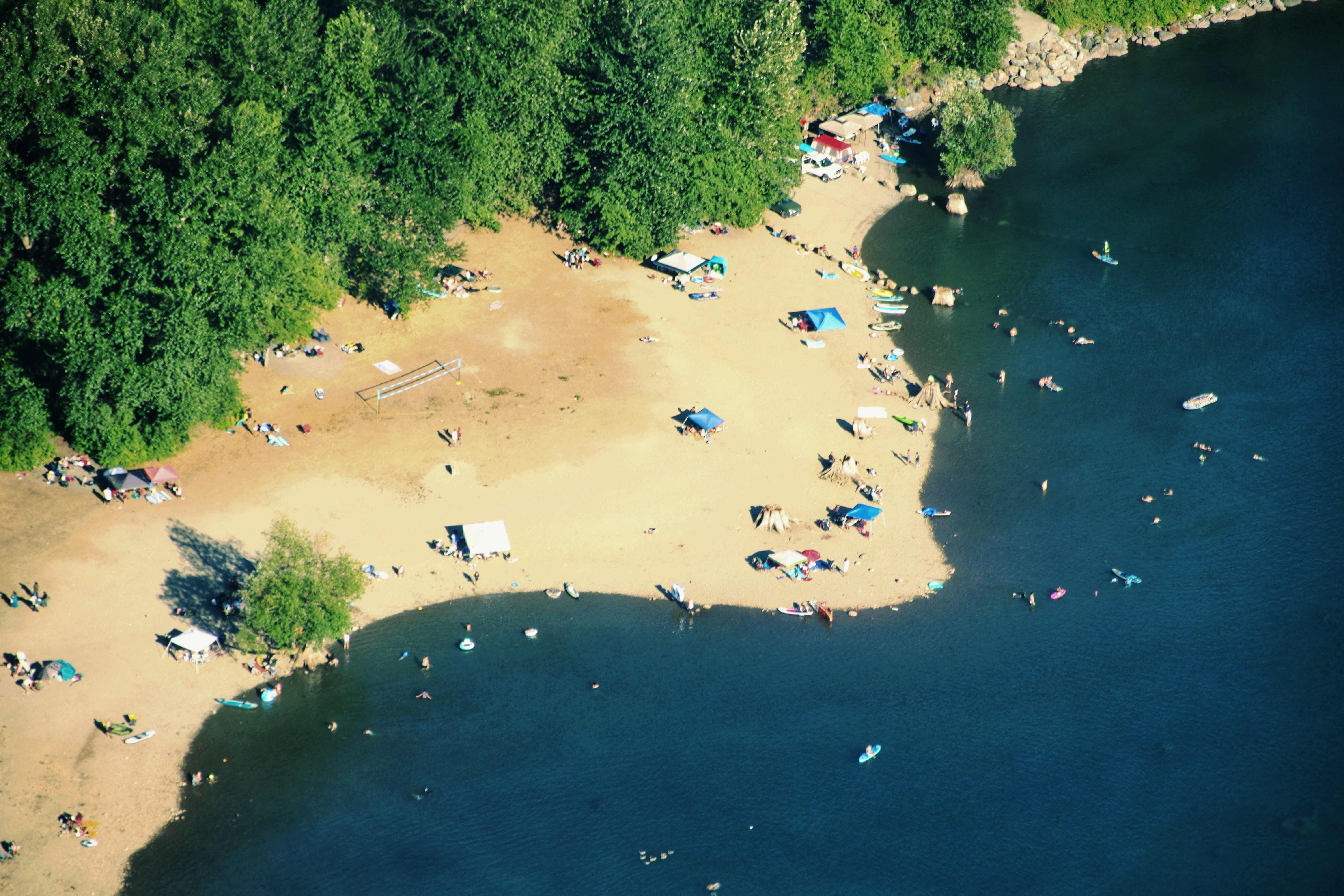 Aerial shot of lake shore populated with day trippers, with navy water, golden sand, and green trees at background