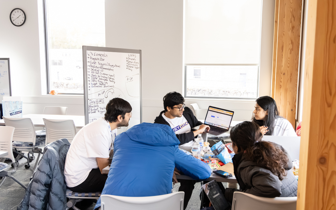 Five students surrounding a laptop and looking at the screen together as they brainstorm in front of a whiteboard.