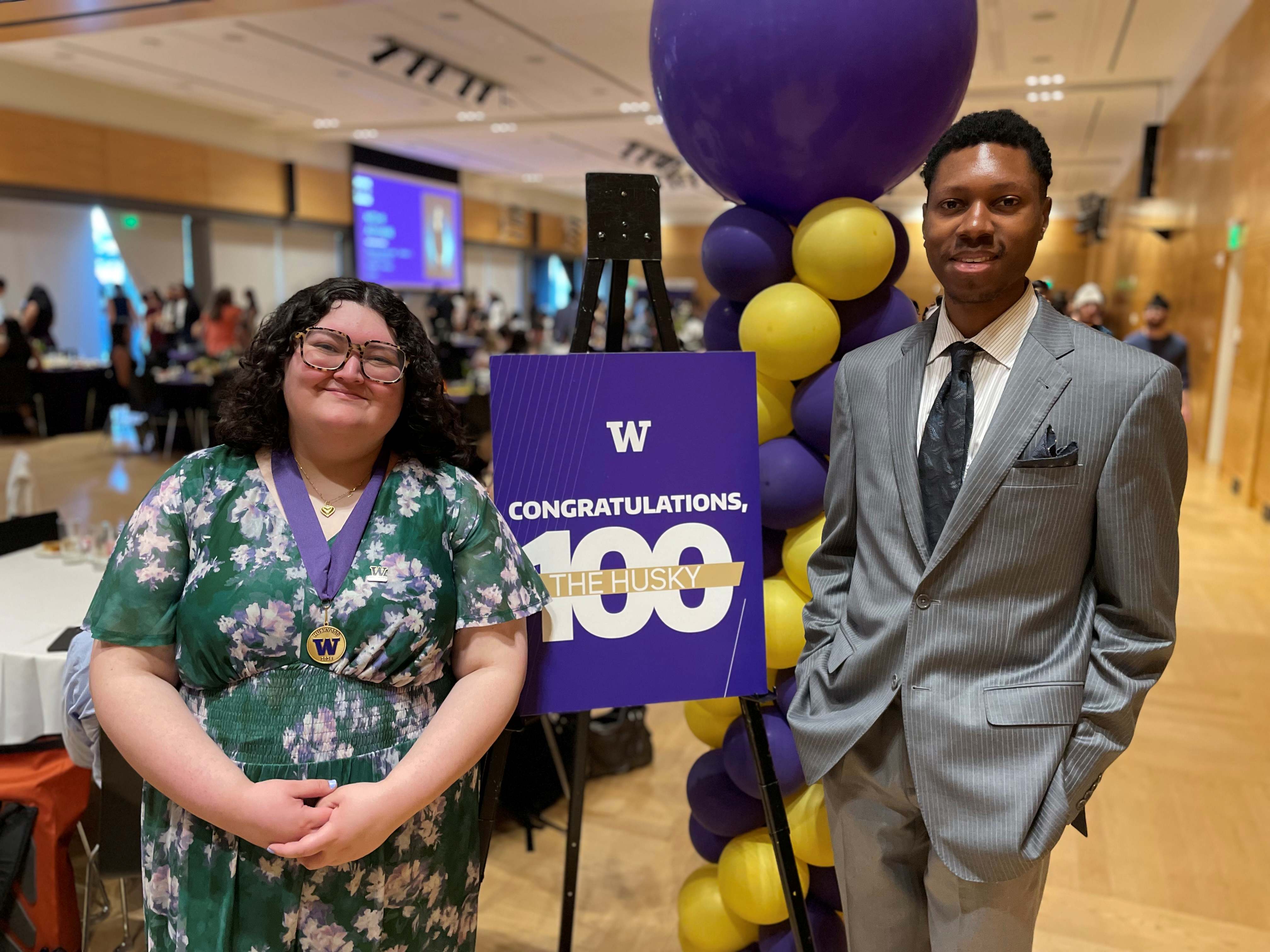 Two people standing side by side at an indoor event hall, posing in front of a purple sign on an easel that reads “W Congratulations, 100 The Husky.” The person on the left wears a green floral dress and a medal on a ribbon, while the person on the right wears a gray suit with a tie and pocket square. Behind them is a column of purple and yellow balloons. The background shows a banquet-style setting with tables, people mingling, and a large screen with a presentation.