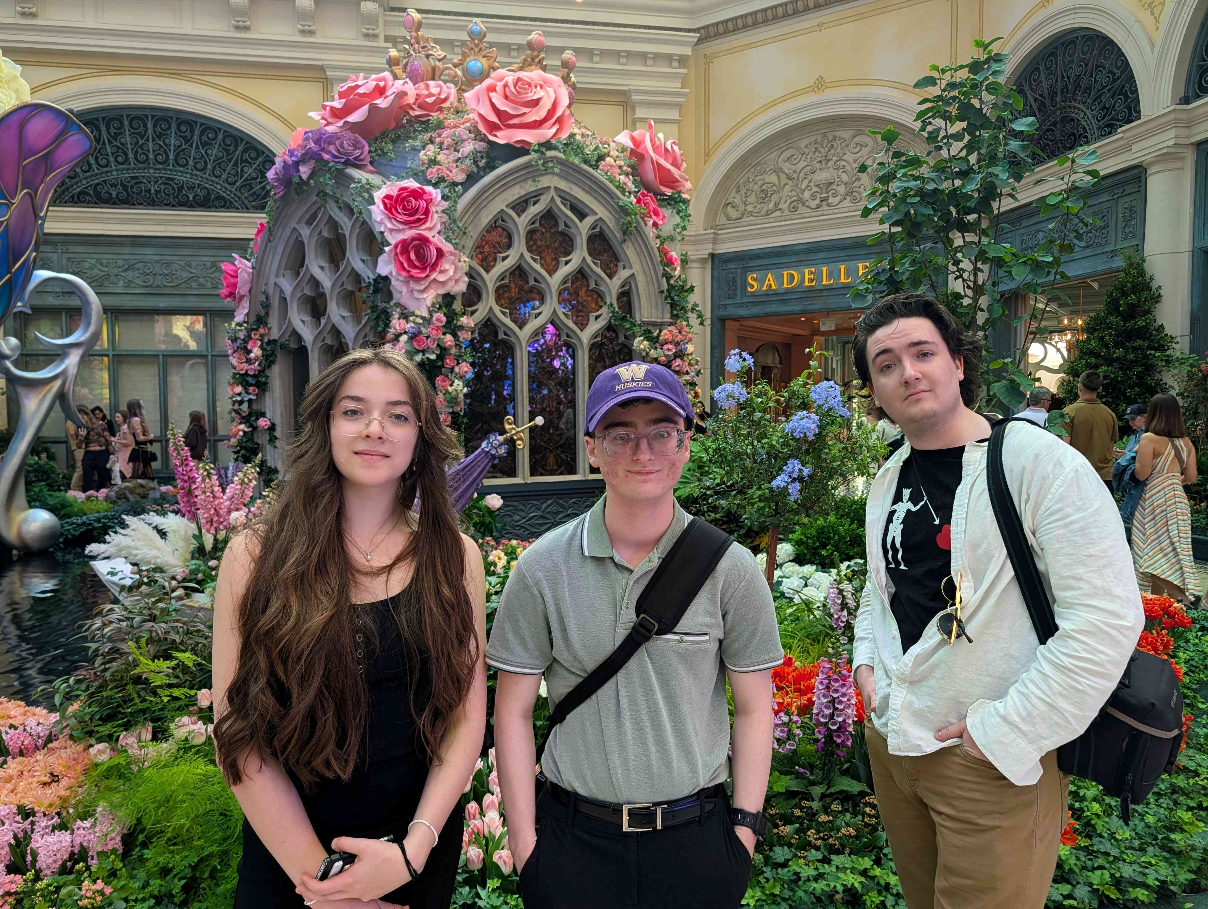 Three people stand in a flower-filled indoor garden in front of a rose-covered decorative structure with crown details; a “SADELLE’S” sign is visible in the background.