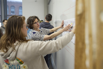 Students stand at a large whiteboard covered with a grid, placing and organizing notes as part of a collaborative project in a classroom or workshop setting.