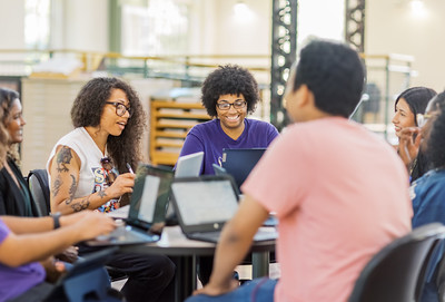 Students sit around a table using laptops and discussing ideas in a collaborative learning space on campus.