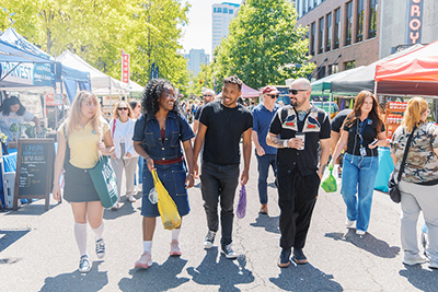 UW Tacoma students stroll through an outdoor farmers market in downtown Tacoma, with vendor booths, local produce, and tree-lined city streets visible in the background.