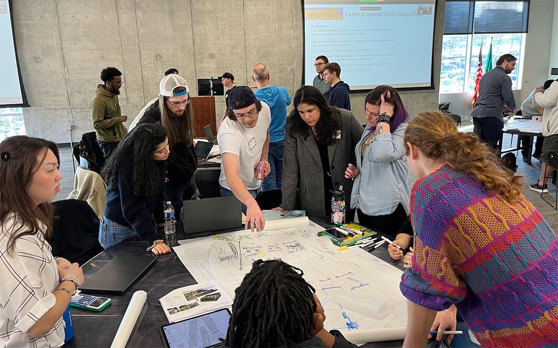 Group of participants standing around a table, reviewing and marking up a large paper map with pens and notes during a collaborative design workshop in a classroom setting.