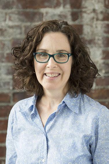 Head-and-shoulders portrait of a person with shoulder-length curly hair and teal-framed glasses, wearing a light blue patterned button-up shirt. The person is facing the camera against a textured brick wall background with soft, even lighting.