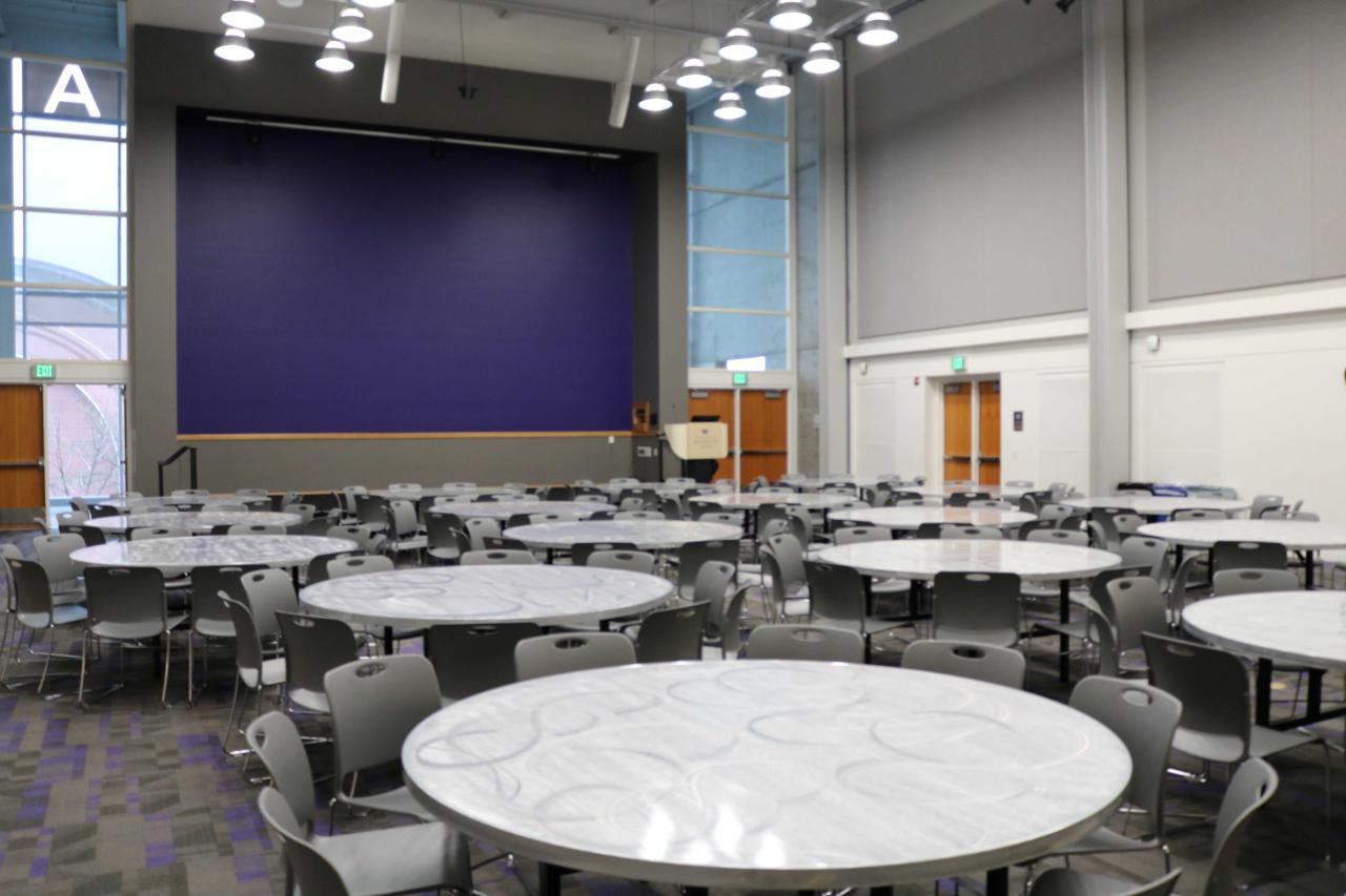 Event space full of round tables with chairs and a stage with a purple wall behind it.