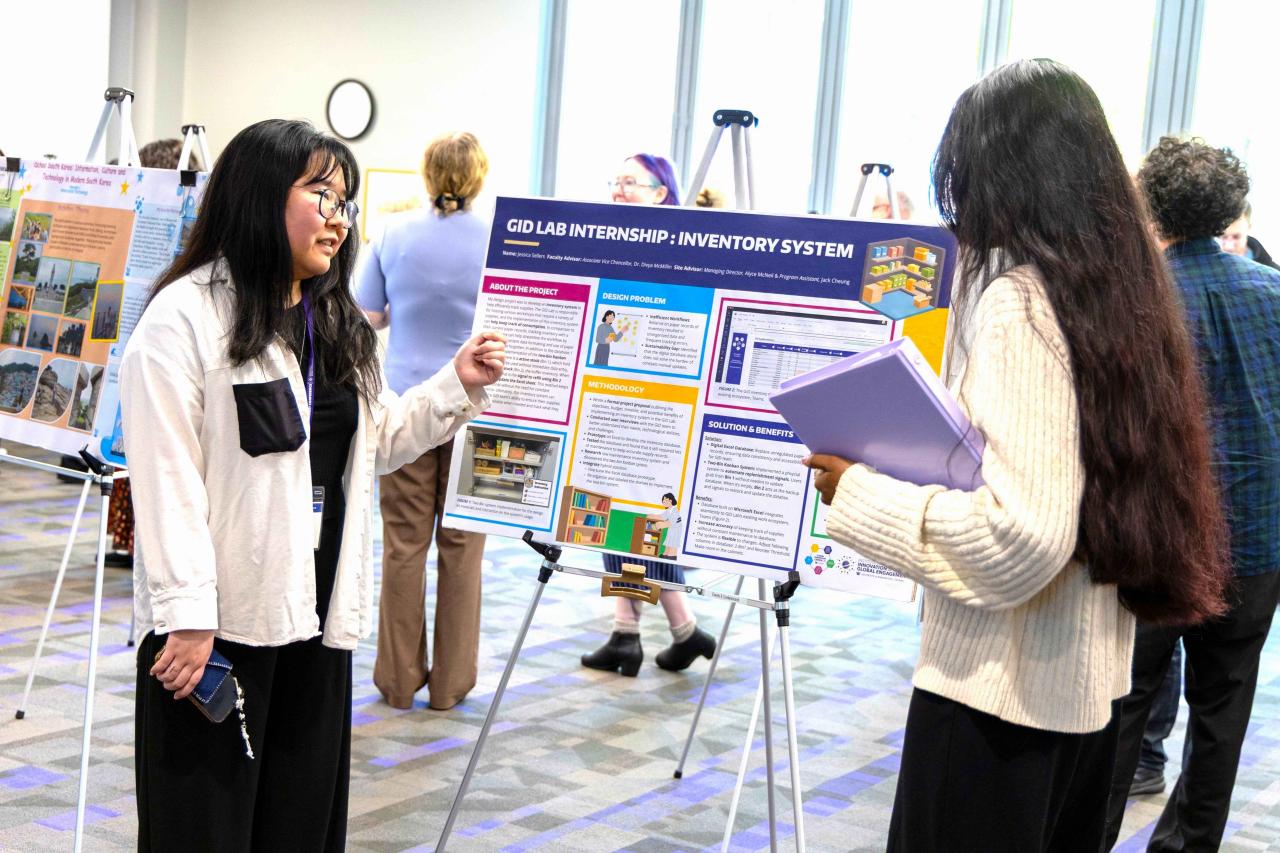 Attendee presents a poster titled “GID Lab Internship: Inventory System” during a research poster session, pointing to project details while speaking with another attendee holding a folder. Multiple poster displays and participants are visible in a bright conference space with large windows.
