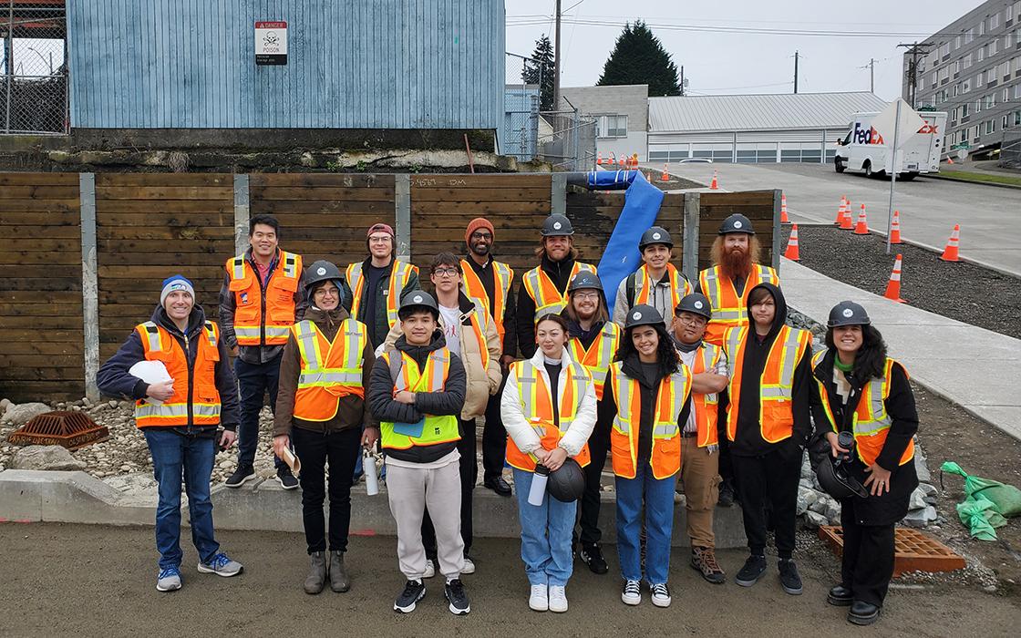 Group of participants wearing orange safety vests and hard hats pose together at an outdoor construction site, with traffic cones, a retaining wall, and nearby buildings in the background.