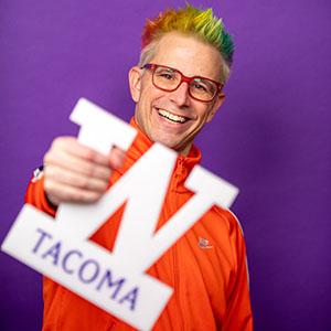 UW Tacoma senior Alex Zerbe is holding a small, plastic version of the steel W. He is smiling and has green hair and an orange jacket.