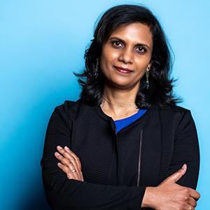 UW Tacoma faculty member Menaka Abraham stands in front of a blue background. Her arms are crossed in front of her. She is wearing a black coat with a blue blouse underneath.