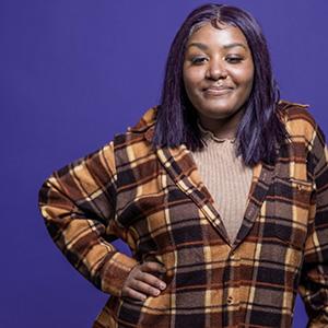 UW Tacoma student Chanise Jackson stands against a purple background. She is wearing a plaid shirt and is holding onto a walker.