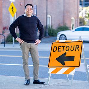 UW Tacoma faculty member Ruben Casas stands next to an orange detour sign. The arrow on the sign is pointing to the left. Casas is wearing khaki pants and a black shirt. There is a street in the background.