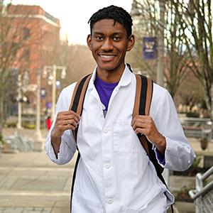 UW Tacoma junior Christian James stands on the grand staircase. He has a backpack on his shoulders. James is wearing a white lab coat with a purple shirt underneath. There are campus buildings and trees in the background.