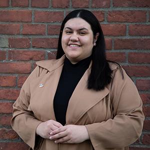 Headshot of UW Tacoma student Exita Lealofi. She is wearing a long, brown trench coat and has long, black hair. Lealofi is standing in front of a red brick wall.
