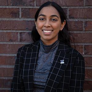 UW Tacoma student Angel Reddy stands against a red brick wall. She is wearing a red, checkered coat with a gray undershirt. There is a purple and gold UW Tacoma pen on her lapel. Reddy is smiling and has long black hair.