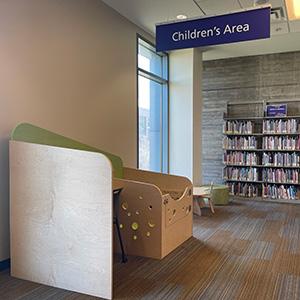 Picture of wood study carrel which resembles a desk. There is a sign that reads "Children's Area" in the background. There are books on shelves and a toy in the background.