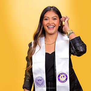 UW Tacoma alumna Victoria Nuon stands against a gold background. Her left hand is near her ear like she's moving a stand of hair. Nuon has long brown hair and has a white UW Tacoma sash around her neck. She is wearing a dark colored blouse.
