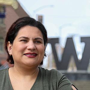 Close up of recent Master of Nursing graduate Juana Gallegos. She has short, dark hair and is wearing a brown top. In the background are the steel "W" on campus and part of a red brick building.