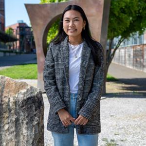 UW Tacoma alumna Teiya Shimomura stands in front of Maru on campus. Maru is a bronze sculpture that looks like a square with a hole in the middle. Shimomura has long, dark hair and is wearing a plaid jacket, white undershirt and jeans. There is grass and a wood wall in the background.