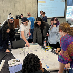 Group of participants standing around a table, reviewing and marking up a large paper map with pens and notes during a collaborative design workshop in a classroom setting.