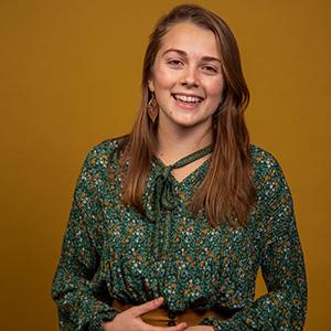 UW Tacoma alumna Abigail Lawson stands in front of a gold colored background. She has long brown hair and is wearing a dark, floral print shirt.