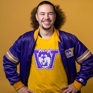 UW Tacoma student Jai'Shon Berry stands. His arms are on his hips. He is wearing a gold UW undershirt that features a husky and a "W." Berry is also wearing a purple UW jacket. Berry's dark hair is pulled back in a ponytail.