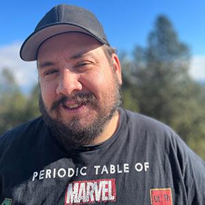 Alumnus Steve Stermer. He is wearing a dark colored hat and a black Marvel t-shirt that reads "Periodic Table of Marvel." He has a beard. There are trees and blue sky in the background.