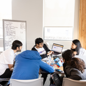 Five students surrounding a laptop and looking at the screen together to brainstorm.