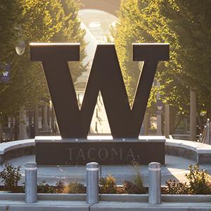 The University of Washington Tacoma “W” statue in the center of the UW Tacoma campus. Tree-lined walkways and campus buildings extend into the background in warm sunlight.