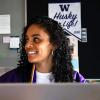 UW Tacoma alumna Amalia Perez sits at a desk. In the background is a poster that says "Husky for Life" in purple letters. Perez has long, black, curly hair and is wearing a purple and gold jacket with a white sweater underneath.