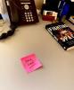 Handwritten message reading "Back Soon" on pink post-it note lying on desk near telephone and book.