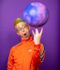 Alex Zerbe, wearing orange jacket and with rainbow-hued hair, balances a large ball on his W-configured fingers.
