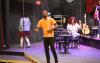 A man stands on the stage at the Tacoma Little Theatre. He has short black hair and is wearing a gold colored shirt.